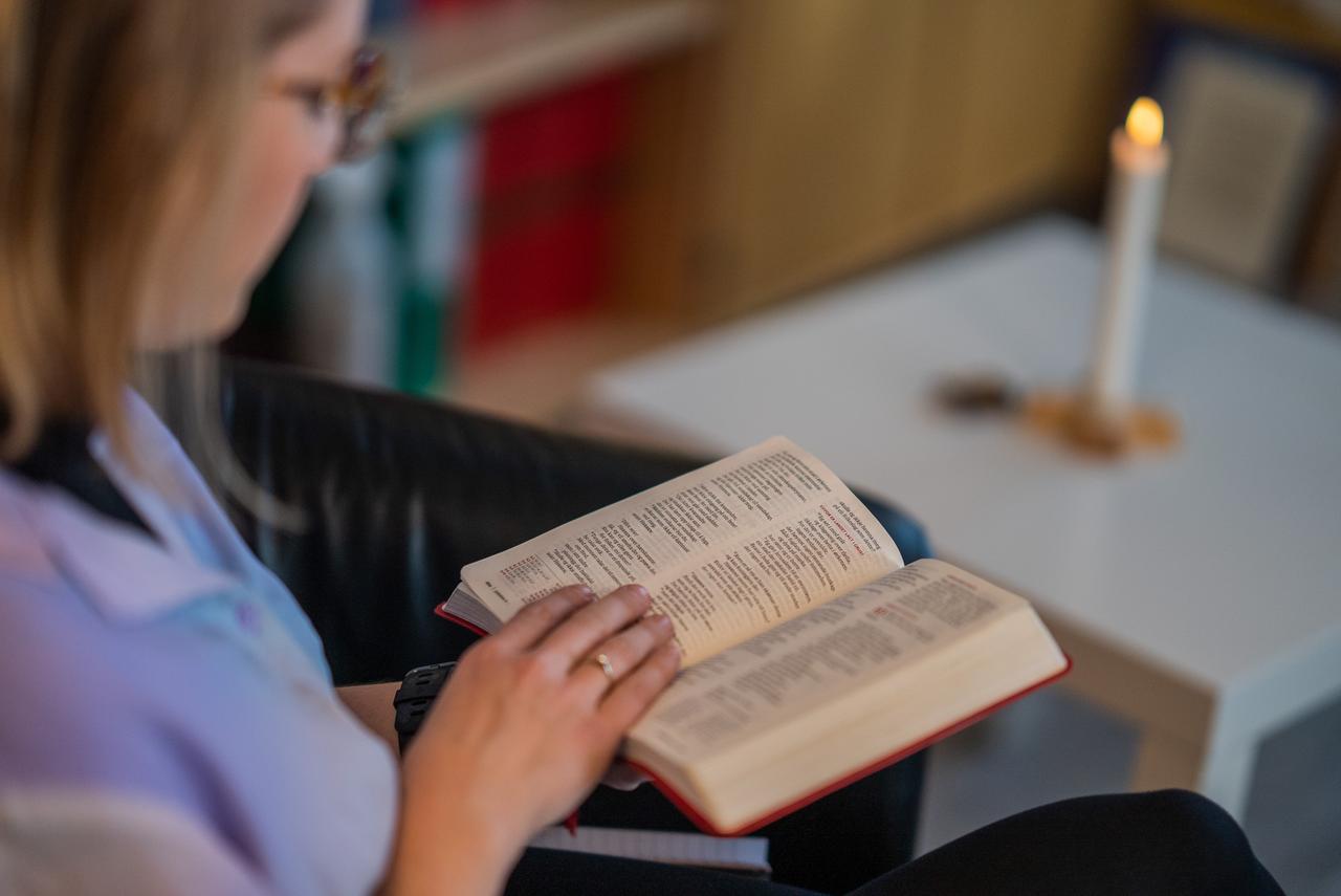 Kvinne sitter og blar i en bibel, ved et bord med tent lys.